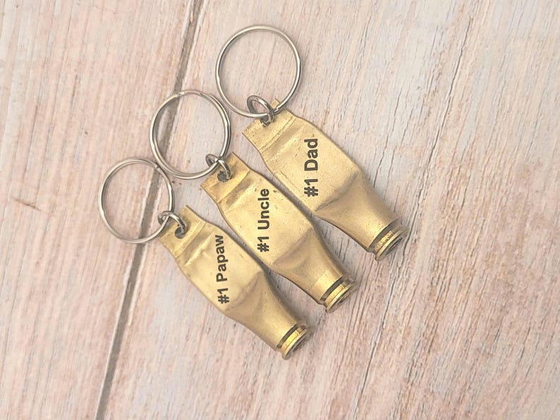 Puede incluir: Tres llaveros de color dorado con texto grabado. Los llaveros tienen forma de casquillos de bala y tienen el texto "#1 Papaw", "#1 Uncle" y "#1 Dad" grabado en ellos.