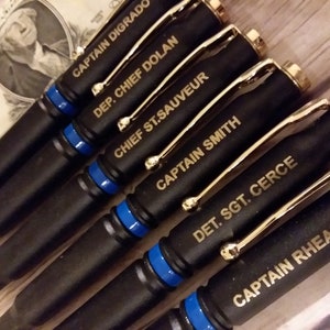 Artful 45° angle picture of engraved thin Blue line bullet pens for police officers. The pens are flat black with gold components and a blue line separator with gold engraving on the top bullet