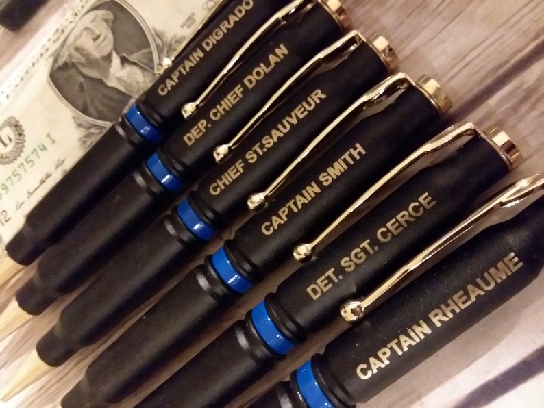 Artful 45° angle picture of engraved thin Blue line bullet pens for police officers. The pens are flat black with gold components and a blue line separator with gold engraving on the top bullet
