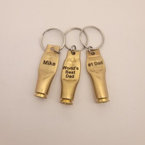 Puede incluir: Tres llaveros de color dorado con texto grabado. Los llaveros tienen forma de casquillos de bala. El texto en los llaveros dice "Mike", "World's Best Dad" y "#1 Dad".