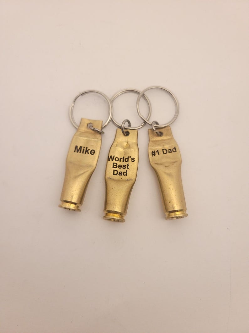 Puede incluir: Tres llaveros de color dorado con texto grabado. Los llaveros tienen forma de casquillos de bala. El texto en los llaveros dice "Mike", "World's Best Dad" y "#1 Dad".