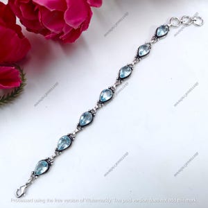 Peut inclure: Un bracelet en argent orné de sept pierres précieuses bleues claires en forme de poire. Le bracelet est relié par une série de petits maillons décoratifs. Le fermoir est un simple crochet et œillet. Fond blanc avec une fleur rose.