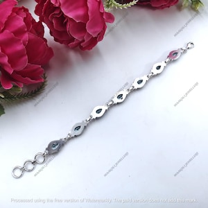 May include: A silver bracelet featuring oval-shaped, light blue gemstones. The bracelet is composed of multiple gemstone settings connected by small links. The gemstones are set in a silver metal. The bracelet is displayed on a white surface with pink flowers in the background.