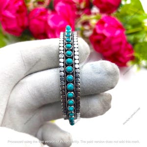 May include: A silver cuff bracelet with turquoise stones. The bracelet features a row of round turquoise stones set in a silver band, with a dotted and patterned border. The background includes pink flowers.
