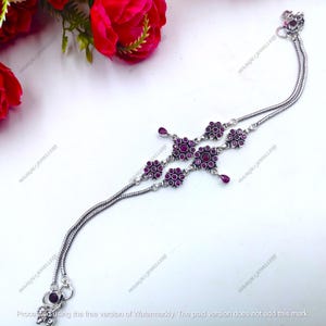 May include: A silver anklet featuring a double snake chain design adorned with multiple floral-shaped clusters set with deep pink stones and small teardrop-shaped dangles. The piece includes a secure clasp and small decorative charms at the ends.