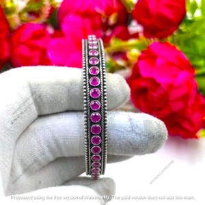 May include: A silver-toned bangle bracelet featuring a single vertical row of round, vibrant pink stones set in a beaded metal frame. The bracelet has a textured, rope-like border on both sides of the central stone setting, creating a detailed design.