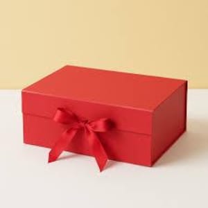 May include: A red rectangular gift box with a matching ribbon tied in a bow. The box is closed and sits on a white surface against a light yellow background. The box is made of a matte material.