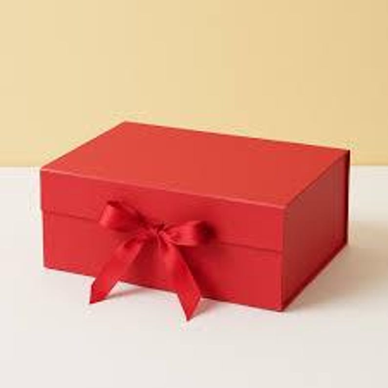 May include: A red rectangular gift box with a matching ribbon tied in a bow. The box is closed and sits on a white surface against a light yellow background. The box is made of a matte material.