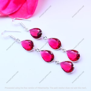 May include: Silver drop earrings with three ruby-red, teardrop-shaped gemstones. The earrings have a hook fastening and are displayed on a white surface. The gemstones are connected by small silver links. The earrings are a jewellery item.