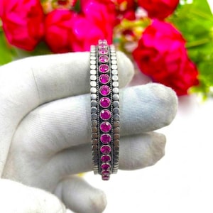 May include: A silver-toned metal bangle bracelet featuring a central row of round, vibrant pink faceted stones. The bracelet is flanked by two rows of small, textured circular metal beads, creating a detailed and structured design.