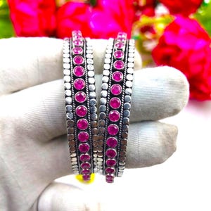 May include: Two silver-toned bangles featuring a central row of round, vibrant pink stones. Each bangle is bordered by textured, beaded metal edges, creating a distinct pattern. The design showcases a metallic finish with bright pink accents.