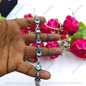 May include: A silver bracelet featuring a series of teardrop-shaped, light blue gemstones. The bracelet is held against a backdrop of pink roses and green foliage, showcasing the jewelry's design and color.