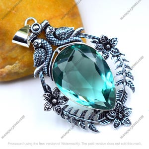 May include: A silver pendant featuring a large, teardrop-shaped, teal-colored gemstone. The pendant is adorned with floral and leaf details, along with two bird figures at the top. The words "MAAKALI JEWELLERS" are printed on the image.
