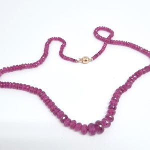 Op de afbeelding: Een ketting bestaande uit gefacetteerde, dieproze edelstenen. De kralen variëren in grootte, met grotere stenen in het midden. Een goudkleurige sluiting zet de ketting vast. De sieraden worden tegen een witte achtergrond getoond.