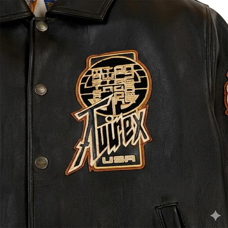 Puede incluir: Chaqueta de cuero negra con cierre de botones y cuello. La chaqueta presenta un gran parche bordado con la palabra "Avirex" y "USA" en una fuente estilizada. El parche es en tonos beige, negro y dorado.