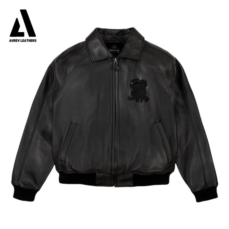 Puede incluir: Chaqueta bomber de cuero negro con cremallera, cuello y pu&ntilde;os y dobladillo de canal&eacute; negros. Cuenta con un emblema negro en el pecho y el logotipo "AUREV LEATHERS" en la esquina superior izquierda.