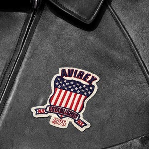 May include: Close-up of a black leather jacket with a patch. The patch is a shield design with red and white stripes, blue stars, and the word "AVIREX". The patch also includes the text "NY ESTABLISHED NY 1975".