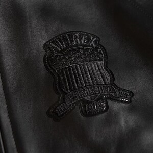 Puede incluir: Primer plano de una chaqueta de cuero negra con una superficie texturizada. Un emblema negro est&aacute; cosido en la chaqueta, con el texto "AVIREX" y "ESTABLISHED 1975 NYT". El emblema tiene forma de escudo con l&iacute;neas verticales.