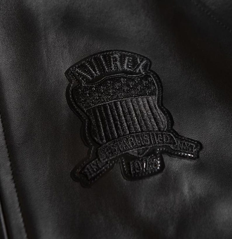 Puede incluir: Primer plano de una chaqueta de cuero negra con una superficie texturizada. Un emblema negro est&aacute; cosido en la chaqueta, con el texto "AVIREX" y "ESTABLISHED 1975 NYT". El emblema tiene forma de escudo con l&iacute;neas verticales.