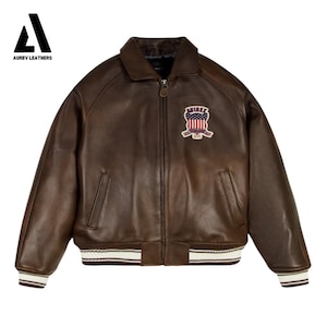 May include: A brown leather bomber jacket with a zip-up front, a collar, and a patch on the chest. The jacket has white and brown striped cuffs and hem. The brand "AUREV LEATHERS" is in the top left corner.