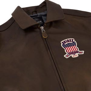 May include: A brown leather jacket with a collar and a zipper closure. The jacket features a patch with the text "AVIREX ESTABLISHED 1975" and a design of the American flag.