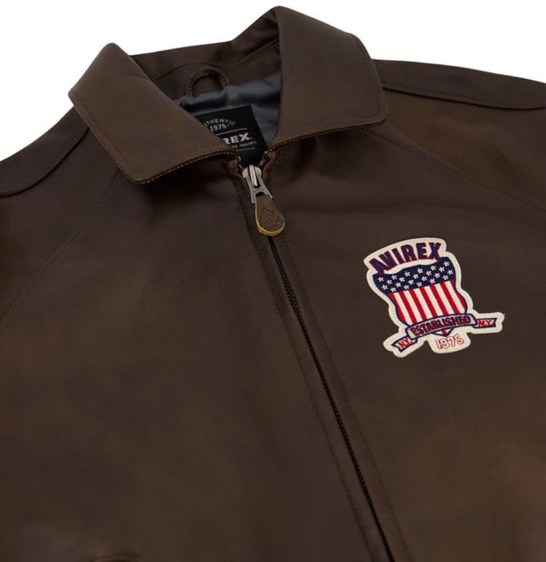 May include: A brown leather jacket with a collar and a zipper closure. The jacket features a patch with the text "AVIREX ESTABLISHED 1975" and a design of the American flag.