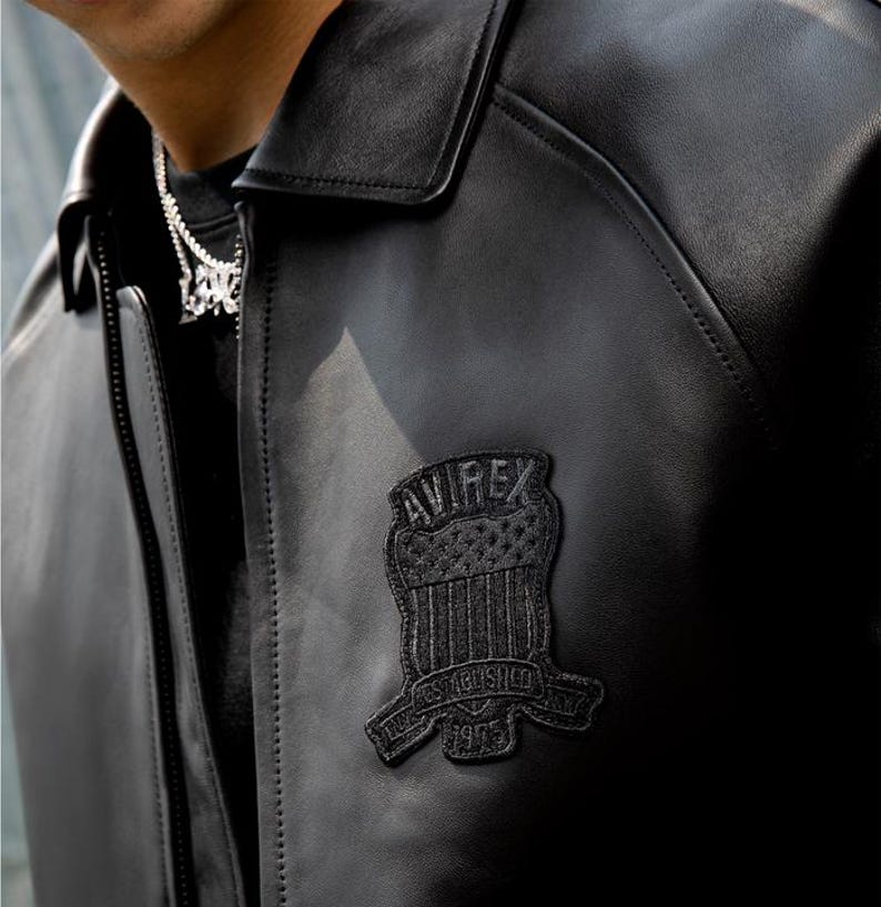 Puede incluir: Chaqueta de cuero negro con cuello y cierre de cremallera. La chaqueta presenta un parche negro en el pecho con el texto "AVIREX" y "ESTABLISHED 1975". Chaqueta de estilo cl&aacute;sico.