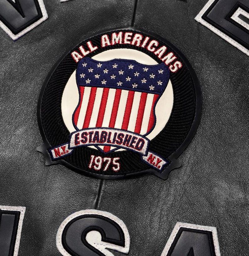 May include: Close-up of a black leather jacket with a circular patch. The patch features the words "ALL AMERICANS" and "ESTABLISHED 1975" surrounding a shield design with the American flag. White lettering is visible.
