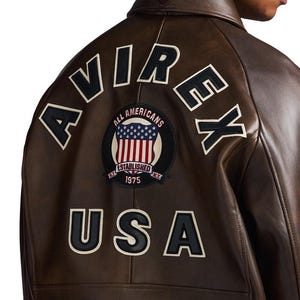 May include: Brown leather jacket with white lettering spelling "AVIREX" and "USA" on the back. A circular patch features the American flag and the words "ALL AMERICANS ESTABLISHED 1975 N.Y."