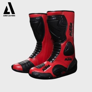 Könnte beinhalten: Paar rote und schwarze Motorradstiefel. Die Stiefel haben ein hoch geschnittenes Design mit schwarzen Schutzpaneelen und dem KTM Racing Team-Logo. Das Aurev Leathers-Logo befindet sich in der oberen linken Ecke.