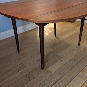 Op de afbeelding: Een ronde houten eettafel met een warme bruine afwerking en een zichtbare houtnerf. De tafel heeft een glad, cirkelvormig tafelblad en vier taps toelopende, donkerbruine poten die vanuit het midden naar buiten toe zijn geplaatst.