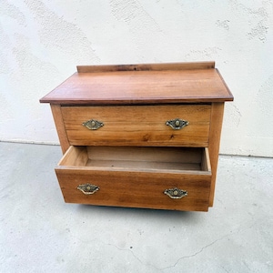 May include: A small wooden chest of drawers featuring a natural light brown wood grain finish, a flat top surface with a back gallery rail, and two drawers. Each drawer is fitted with two ornate, antique-style brass metal pull handles.