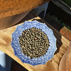 May include: A blue and white decorative plate with a scalloped edge, filled with small, dark-colored stones. The plate sits on a wooden surface, with other pottery visible in the background. The plate's design features a blue floral pattern.