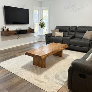 May include: A natural wood coffee table with a live edge design sits on a cream-colored rug in a living room. The table is rectangular with a light brown finish. A dark gray sectional sofa and a wall-mounted TV are also visible.