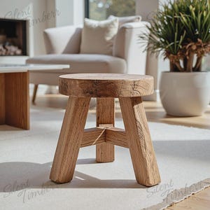 Könnte beinhalten: Ein kleiner, runder Holzschemel mit drei angewinkelten Beinen. Der Hocker ist aus hellbraunem Holz gefertigt und hat ein natürliches, rustikales Aussehen. Der Hocker steht auf einem hellen Teppich in einem hellen Raum.