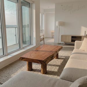 May include: A natural wood coffee table with a live edge design. The table is rectangular with a rich brown color and sits on four cylindrical legs. The table is placed on a neutral-colored rug in a modern living room setting.