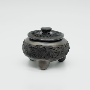May include: A dark gray, handcrafted ceramic jar with a lid. The jar features intricate floral and leaf carvings around the body and on the lid. It stands on three small feet and has a small knob on top of the lid.