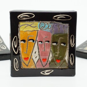 May include: A square, black ceramic tile with a colorful, abstract design of three faces. The faces are in yellow, pink, and green, with red lips and black outlines. White swirls and lines decorate the black border.