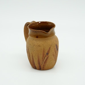 May include: A handmade ceramic pitcher with a handle. The pitcher is a warm brown color with darker brown accents and a textured surface. Vertical, leaf-like patterns decorate the body of the pitcher.