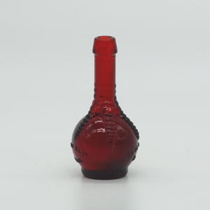 May include: A small, ruby-red glass bottle with a bulbous base and a long, slender neck. The bottle has a textured design around the base and a smooth, rounded top. The bottle is empty and stands upright against a white background.
