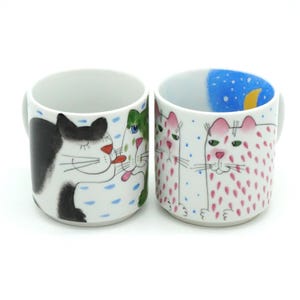 May include: Two ceramic mugs with cat illustrations. One mug features a black and white cat, the other has pink spotted cats. The mugs have white interiors and gray handles. The background of the mugs is white with blue and pink accents.