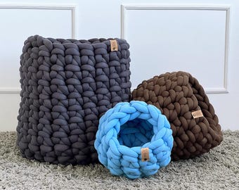 May include: A set of three chunky knit baskets in varying sizes and colors. The largest is a dark gray square basket, a medium-sized brown dome-shaped basket, and a small light blue round basket. Each basket has a leather tag.