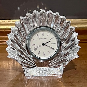 Puede incluir: Un reloj de cristal transparente con un diseño en forma de abanico. La esfera del reloj es blanca con números romanos y manecillas negras. El texto "WATERFORD CRYSTAL QUARTZ" es visible en la esfera. El reloj está sobre una superficie de madera.
