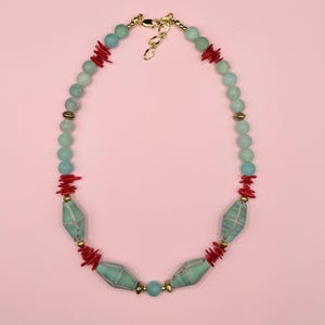 Puede incluir: Un collar con cuentas de color azul claro, rojo y dorado. El collar tiene un cierre dorado e incluye cuentas redondas, piezas rojas con forma de coral y cuentas turquesas más grandes y estampadas. El collar se muestra sobre un fondo rosa.