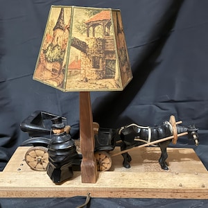 May include: A table lamp featuring a wooden base with a black horse-drawn carriage figurine and a driver figure. A central wooden post supports a hexagonal lampshade decorated with a printed illustration of a stone building and landscape scenery.