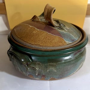 Puede incluir: Una fuente de horno redonda de cerámica con tapa que presenta un asa curva prominente. La base es de un color verde bosque profundo con un esmalte brillante, mientras que la tapa muestra un acabado texturizado y moteado en beige y marrón.