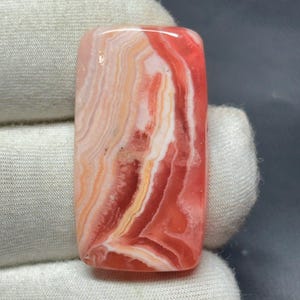 以下が含まれることがあります： 研磨された表面を持つ長方形の宝石。石は赤、白、オレンジの色合いの渦巻き模様を特徴としています。宝石は手袋をはめた手で持たれており、鮮やかな色とユニークなデザインが際立っています。