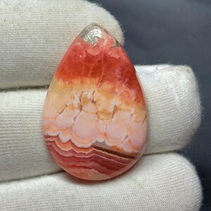 以下が含まれることがあります： 鮮やかな色のグラデーションを持つ、研磨された涙型の宝石。上部は鮮やかな赤色で、オレンジ色に変わり、次にクリーム色、最後に濃い赤褐色の基部で終わります。石の表面は滑らかで、光沢があります。