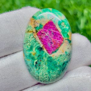 May include: An oval-shaped cabochon gemstone featuring a vibrant magenta-pink diamond-shaped inclusion centred within a mottled surface of bright green, teal, and creamy white mineral patterns with a smooth, polished, and reflective finish.