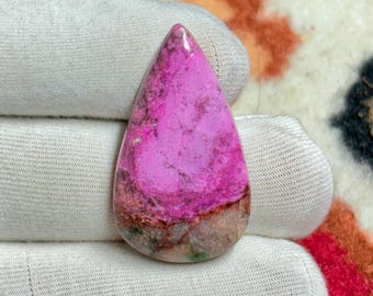 May include: A polished, teardrop-shaped gemstone with a vibrant pink hue, featuring darker pink and brown marbling. The stone is held against a white fabric background, showcasing its color and texture. This gemstone is likely for jewelry making.
